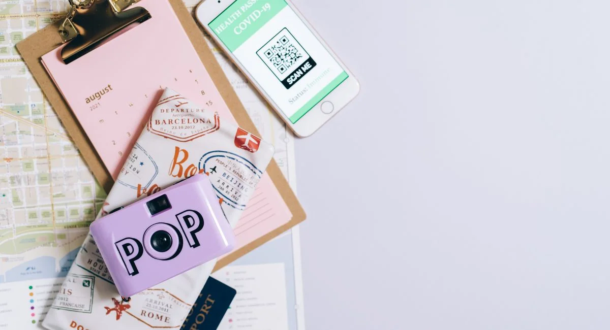 Flat lay of travel essentials including COVID-19 health passport, map, passport, and vintage camera.